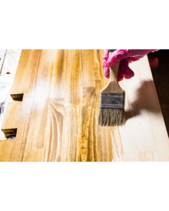A close-up of a hand wearing pink gloves brushing a glossy wood finish onto a light-colored wooden surface, bringing out its texture and shine. CFL Custom Carpentry guarantees the best wood finishes for a stunning and long-lasting result.