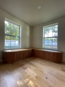 A modern, minimalistic space with natural wood custom banquettes under two large windows, highlighting a sleek design with vertical wood paneling and light hardwood floors.