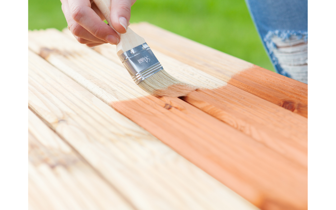 A person outdoors using a paintbrush to apply a protective wood stain on a wooden plank, enhancing its natural beauty. CFL Custom Carpentry delivers the best wood finishes to protect and preserve quality woodwork.