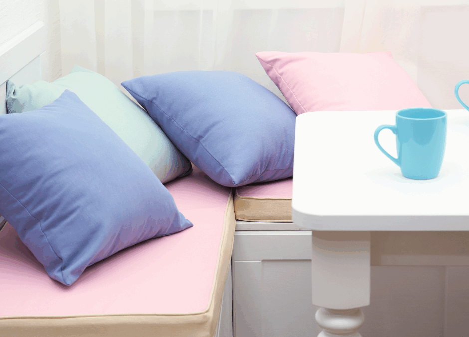 A cozy white corner nook with pastel-colored cushions on a pink bench seat, featuring custom banquettes and a white table with two light blue mugs.